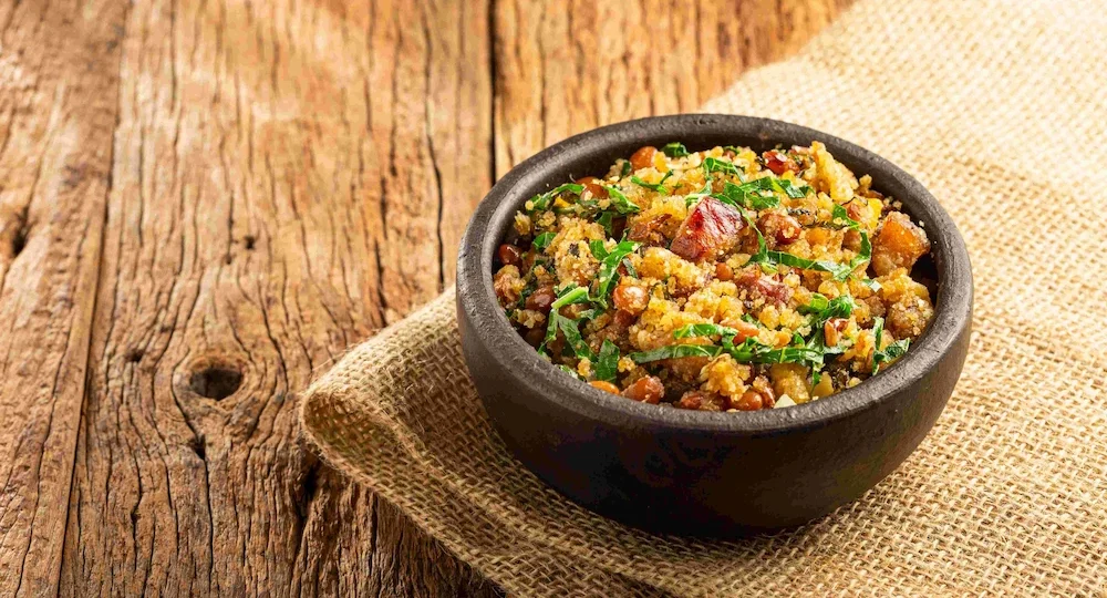 Uma tigela rústica repleta de uma farofa rica e crocante, misturada com grãos de feijão, suculentos pedaços dourados de bacon e tiras finas de couve verde. A tigela está apoiada sobre um tecido rústico de juta, compondo um cenário caseiro, quente e muito apetitoso, típico da culinária brasileira.
