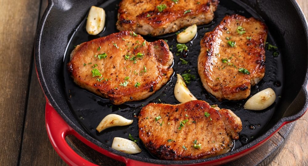 Quatro suculentos cortes de lombo suíno Frimesa com uma crosta perfeitamente dourada e caramelizada, dispostos lado a lado dentro de uma frigideira de ferro fundido. A carne, que brilha com sua própria suculência, é acompanhada por dentes de alho inteiros e finalizada com ervas verdes frescas picadas por cima.