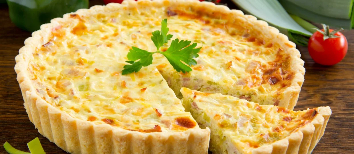 Quiche dourada de bordas onduladas sobre uma mesa rústica de madeira, decorada com um ramo de salsa. Uma fatia cortada revela o recheio cremoso com pedacinhos de frios da Frimesa e vegetais. Ao redor, tomates-cereja, pimentão verde e alho-poró fresco completam a composição.