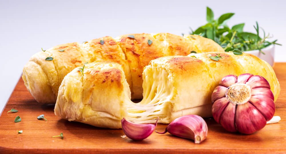 Dois pães de alho dourados e salpicados com ervas finas sobre uma tábua de madeira. O pão da frente está partido ao meio, revelando uma camada generosa de queijo derretido ao esticar entre as metades. Em primeiro plano, ao lado do pão, temos dentes de alho e uma cabeça de alho roxo inteira.