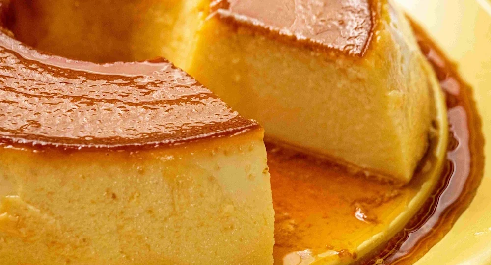 Close apetitoso de um pudim de leite clássico servido em um prato amarelo. O pudim tem uma fatia cortada, revelando um interior incrivelmente liso, cremoso e sem furinhos, indicando que foi assado no ponto perfeito. O topo da sobremesa é coberto por uma calda de caramelo dourada e muito brilhante, que escorre suavemente e se acumula na base do prato.