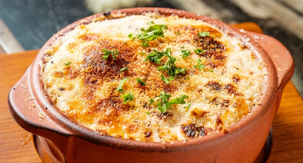Um prato quente e cremoso servido em uma travessa redonda de barro rústico. O preparo exibe uma irresistível cobertura perfeitamente gratinada com queijo Frimesa, formando uma crosta dourada, crocante e com pontos levemente tostados pelo calor, finalizada com ervas verdes frescas picadas por cima. A travessa está apoiada sobre uma tábua de madeira.