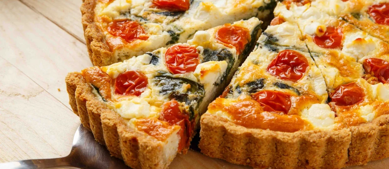 Quiche dourada de bordas onduladas recheada com espinafre, metades de tomates-cereja assados e pedaços de Queijo Frimesa. Uma fatia está sendo levantada por uma espátula de metal, destacando a textura cremosa do recheio e a massa firme. A torta repousa sobre uma mesa de madeira clara sob iluminação quente e convidativa.