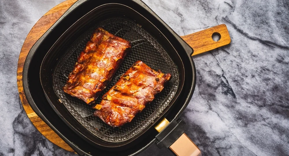 Vista superior de dois pedaços suculentos e bem dourados de Costelinha Suína Frimesa preparados dentro do cesto de uma air fryer. O cesto da fritadeira está apoiado sobre uma tábua redonda de madeira, que repousa em uma superfície com textura de mármore cinza claro.