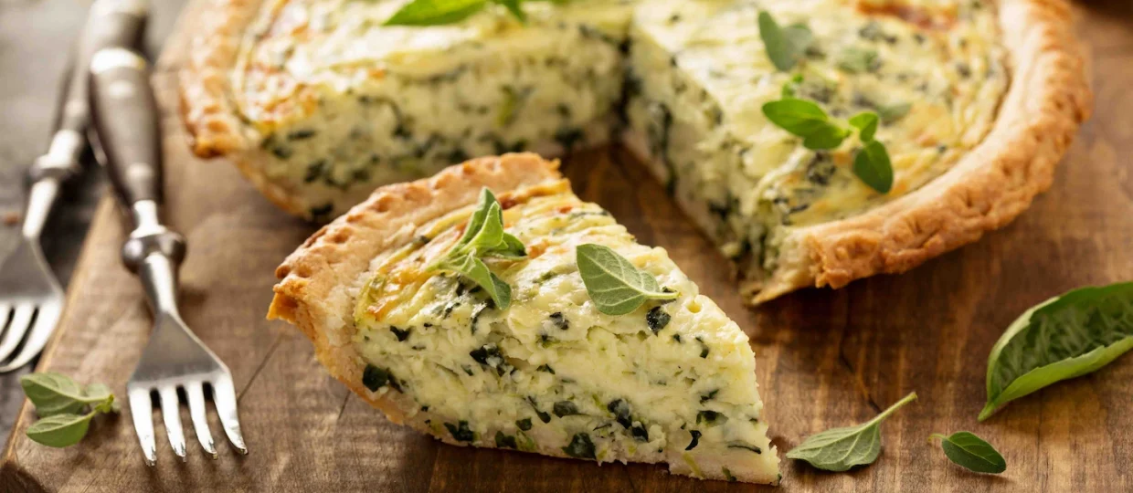 Quiche de Queijo Frimesa com espinafre e ervas de borda dourada servida sobre uma tábua rústica de madeira escura. Uma fatia cortada em destaque revela o recheio denso e cremoso pontilhado de verde, decorada com folhas frescas.
