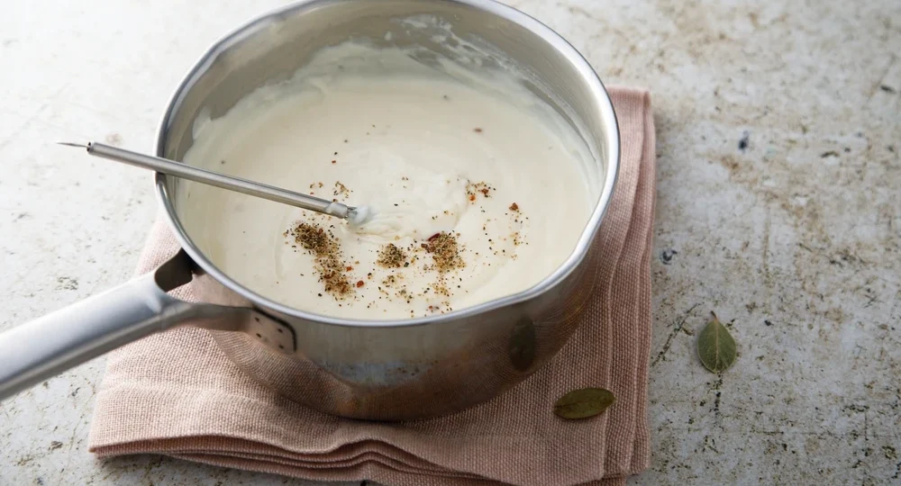 Uma panela de aço inox contendo um molho branco perfeitamente liso, espesso e aveludado, feito com Creme de Leite Frimesa. O molho exibe pimenta moída e especiarias rústicas salpicadas em sua superfície. Um utensílio de metal repousa mergulhado no creme. A panela está apoiada sobre um guardanapo de tecido rosado em cima de uma bancada clara e texturizada, com algumas folhas secas soltas ao redor.