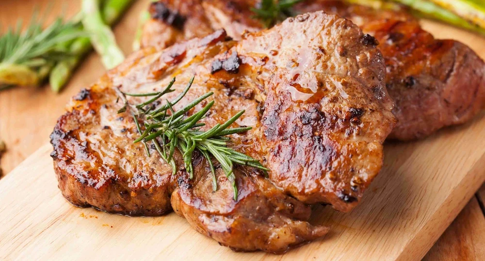 Um close de dois bifes grossos e suculentos de carne suína Frimesa grelhados, servidos sobre uma tábua de corte de madeira clara. A carne apresenta uma crosta bem dourada, brilhante e temperada, decorada com raminhos de alecrim fresco no topo. Ao fundo, levemente desfocados, aspargos verdes acompanham o prato, completando um cenário rústico e apetitoso.