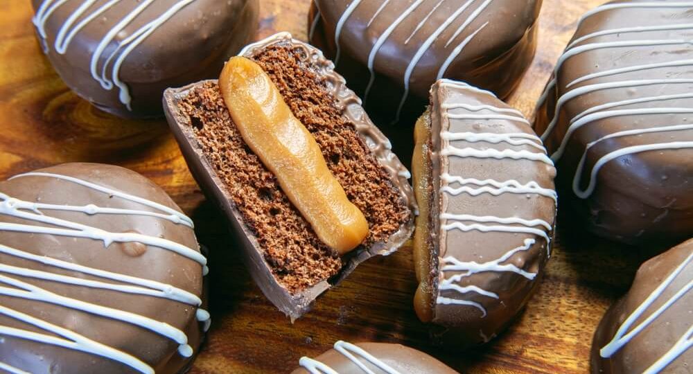 Vários pães de mel redondos cobertos com chocolate de leite e decorados com linhas finas de chocolate branco, dispostos sobre uma superfície de madeira. No centro, um dos pães de mel está cortado ao meio, revelando uma massa fofinha de chocolate com um recheio generoso e cremoso de Doce de Leite Frimesa.