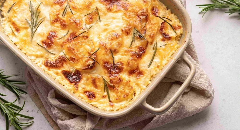 Uma travessa retangular de cerâmica com um apetitoso gratinado de batatas, recém-saído do forno. A superfície exibe uma crosta espessa de Queijo Frimesa derretido e perfeitamente dourada, decorada com finas folhas frescas de alecrim. A travessa repousa sobre um guardanapo de tecido rústico bege, apoiada em uma bancada clara e texturizada, com raminhos extras de alecrim espalhados ao redor, compondo um visual caseiro e irresistível.