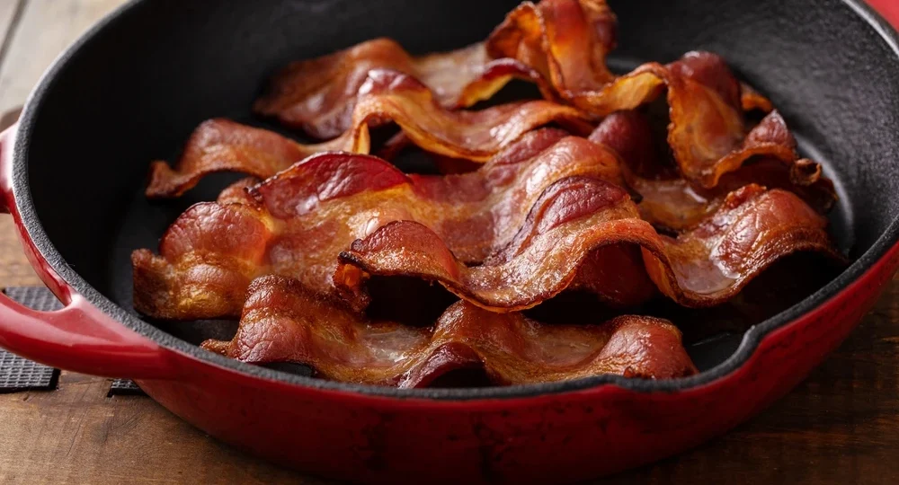 Várias fatias de Bacon Frimesa fritas perfeitamente, douradas e com textura visivelmente crocante, dispostas dentro de uma frigideira de ferro fundido com interior preto e exterior vermelho. A panela está apoiada sobre uma superfície rústica de madeira.