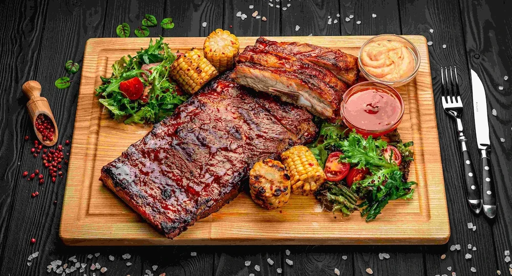 Uma grande e suculenta peça de costelinha suína assada servida sobre uma tábua retangular de madeira clara. A carne está coberta por um molho escuro e brilhante, com uma porção já cortada e empilhada por cima, revelando o interior macio. Acompanham a costelinha pedaços dourados de milho na espiga grelhado, salada verde fresca com tomates-cereja e duas pequenas tigelas de vidro com molhos variados. O prato está sobre uma mesa rústica de madeira escura, decorada com grãos de pimenta rosa, sal grosso e uma pequena concha de madeira rústica à esquerda.