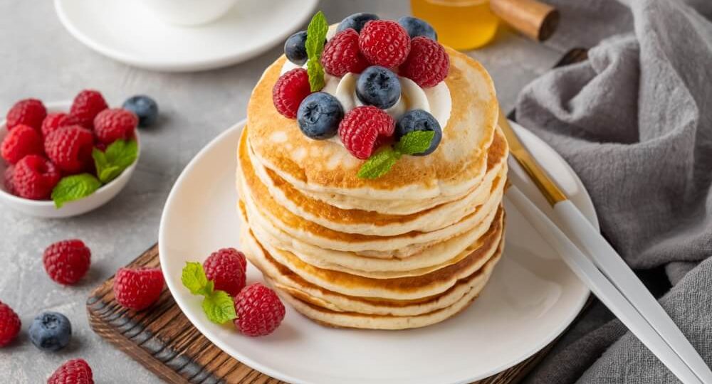 Torre de panquecas americanas fofinhas empilhadas num prato branco, decoradas no topo com uma porção de creme branco, framboesas vermelhas, mirtilos azuis e pequenas folhas de hortelã fresca. Ao lado do prato, sobre a mesa, encontram-se talheres, mais frutos vermelhos espalhados, uma taça com framboesas e um pequeno frasco com mel ao fundo.