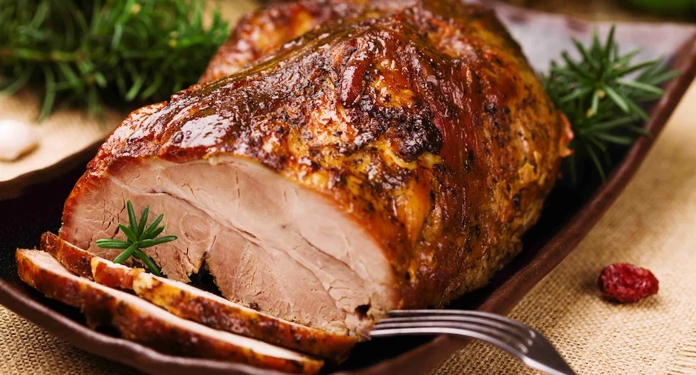 Uma suculenta peça de carne suína assada servida em uma travessa escura. A carne possui uma crosta brilhante, bem dourada e rica em temperos, contrastando com o interior claro e macio revelado pelas primeiras fatias já cortadas na frente. O prato é decorado com pequenos ramos de alecrim fresco, e um garfo de metal repousa elegantemente ao lado das fatias.