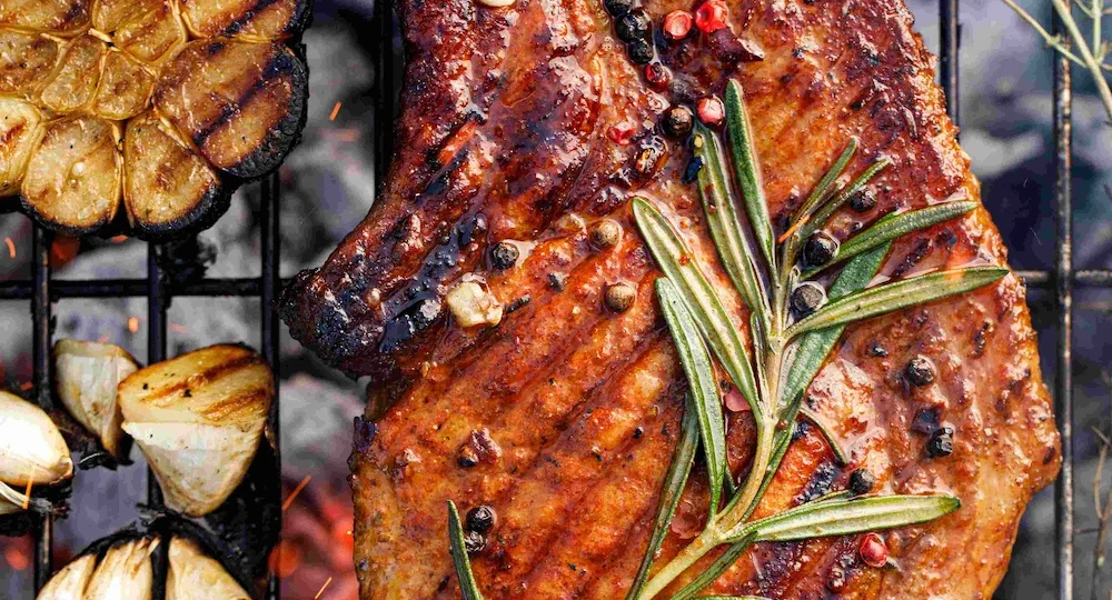Um apetitoso corte suíno com osso assando sobre as grades de uma churrasqueira. A carne exibe uma crosta dourada e suculenta com marcas de grelha, temperada com grãos de pimenta e decorada com um grande ramo de alecrim fresco no topo. Ao redor da peça, grelham-se também uma cabeça de alho cortada ao meio, dentes de alho soltos e pimentas vermelhas inteiras, envoltas por chamas vivas e faíscas que sobem das brasas quentes ao fundo, compondo uma cena de churrasco vibrante e rústica.