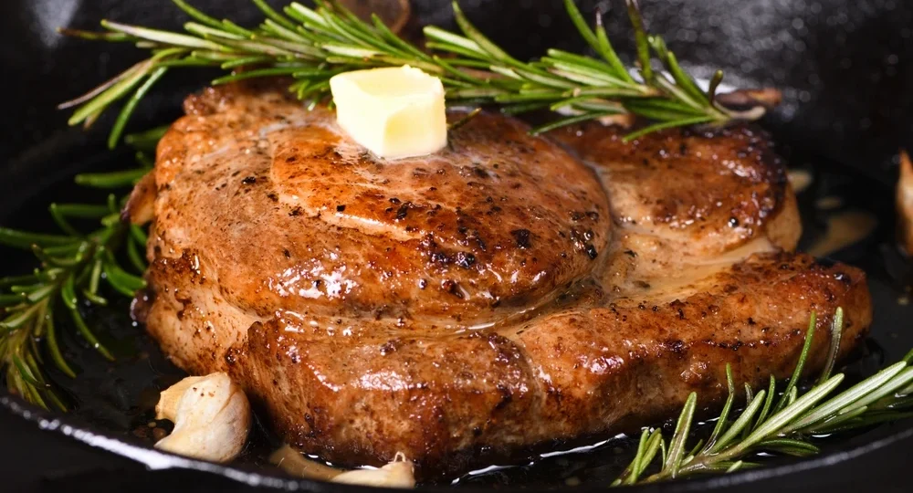 Um corte espesso, macio e suculento de carne suína Frimesa, recém-preparado em uma frigideira escura. A carne exibe uma crosta dourada, brilhante e salpicada com pimenta e temperos rústicos. Bem no centro e sobre a peça quente, repousa um cubo de Manteiga Frimesa derretendo lentamente para trazer ainda mais sabor e suculência ao prato, acompanhado por um ramo de alecrim verde e fresco logo atrás.