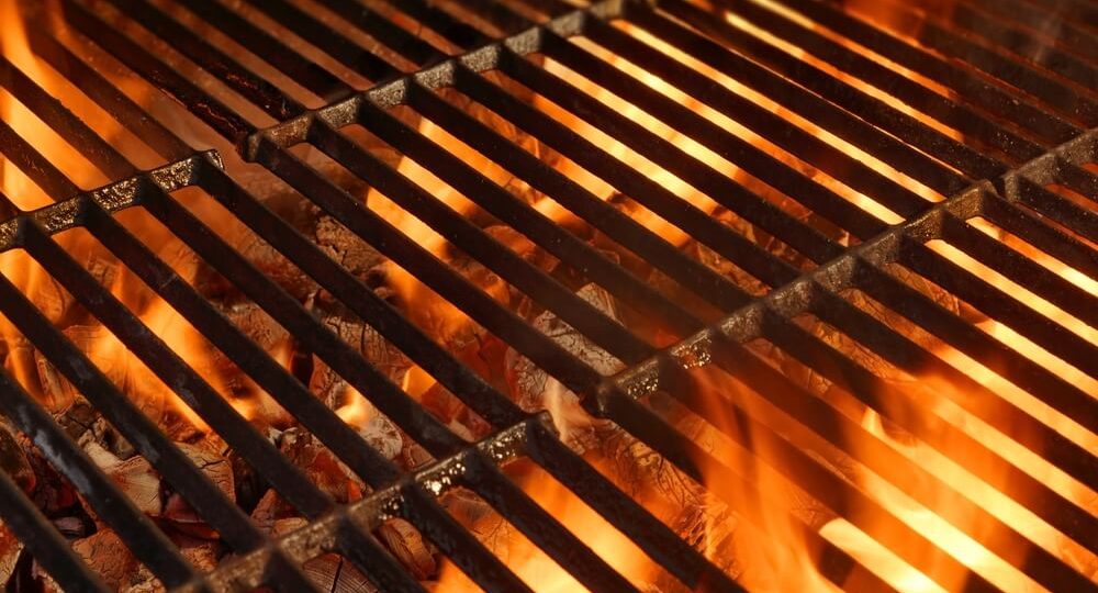 Grelha metálica de churrasco vista de cima, com pedaços de carvão em brasa incandescente no fundo. O carvão em chamas liberta um brilho alaranjado e avermelhado intenso, com chamas quentes que sobem pela grelha.