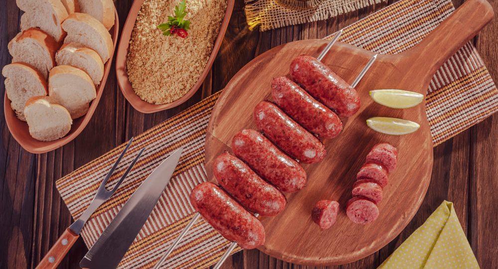Vista superior de suculentas Linguiças Frimesa assadas e douradinhas, servidas em um espeto duplo de metal e apoiadas sobre uma tábua de corte de madeira. Ao lado, uma das linguiças aparece fatiada em rodelas, revelando seu interior perfeitamente cozido e úmido. O cenário de churrasco é completado por fatias de limão, uma faca, panos texturizados e uma tigela com farofa no canto superior esquerdo.