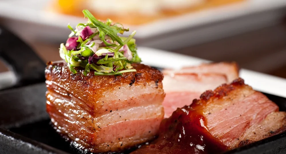 Um espesso e suculento pedaço de Panceta Frimesa servido em uma pequena travessa preta de ferro fundido. A carne exibe camadas perfeitas de gordura e carne rosada, destacando-se por uma crosta superior dourada e bem crocante. Sobre a panceta repousa uma delicada guarnição de folhas verdes e finos pedaços de repolho roxo. Ao lado, outra fatia da carne é acompanhada por um molho escuro, denso e brilhante, compondo um prato sofisticado e extremamente apetitoso com o fundo levemente desfocado.