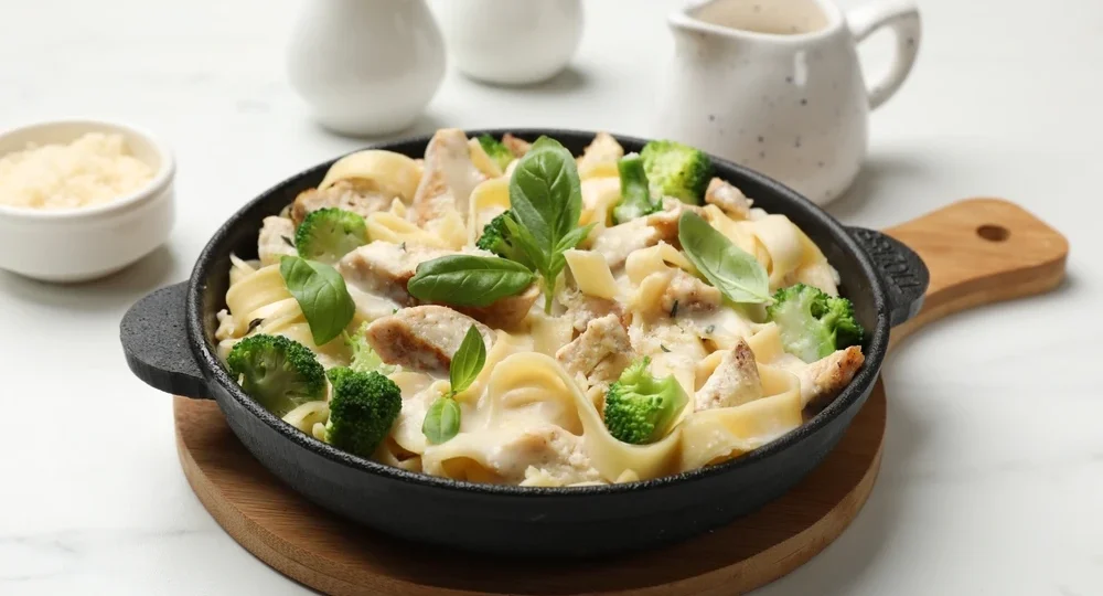 Uma frigideira de ferro fundido preta, apoiada sobre uma tábua redonda de madeira, apresentando uma deliciosa massa tipo fettuccine. O prato é farto e envolto em um molho Alfredo espesso e supercremoso, feito com os queijos Frimesa, misturado a pedaços suculentos de frango grelhado e flores de brócolis de um verde vibrante. No topo, folhas frescas de manjericão decoram e finalizam a receita. O cenário repousa sobre uma bancada clara de mármore, com pequenos utensílios brancos de cerâmica desfocados ao fundo, incluindo uma molheira e uma tigelinha com queijo parmesão ralado.