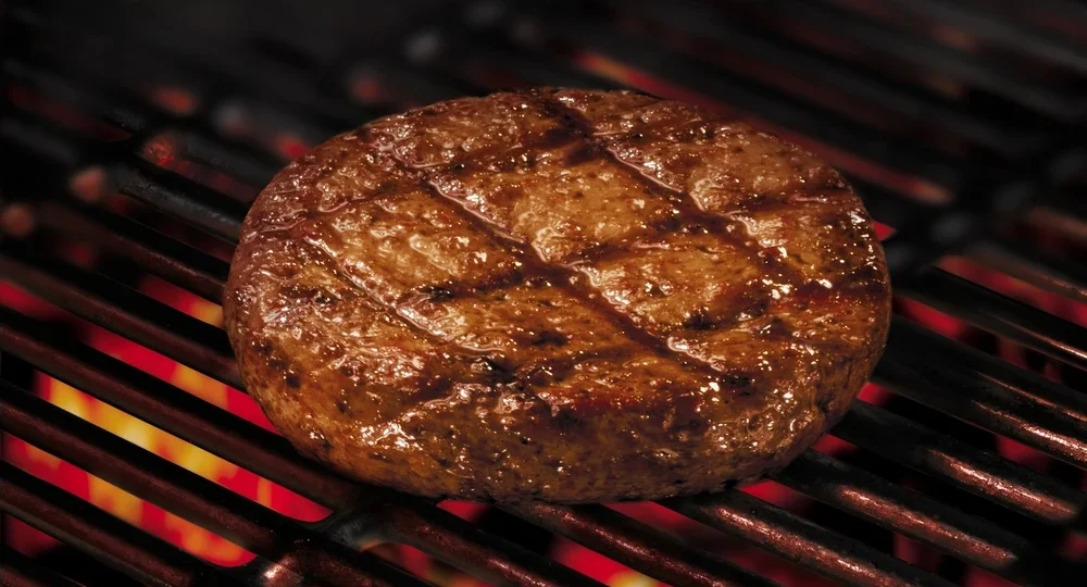 Hambúrguer Frimesa espesso e suculento grelhando em uma churrasqueira sobre chamas vivas. A carne apresenta marcas de grelha bem definidas em formato xadrez e uma superfície brilhante, mantendo seu formato redondo e alto perfeito, sem encolher ou ressecar.