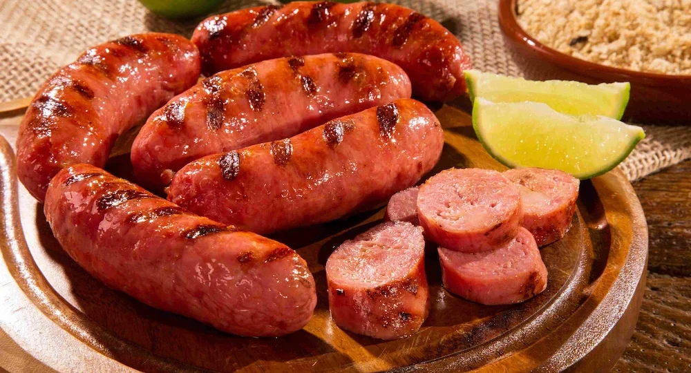 Close apetitoso de Linguiças Frimesa grelhadas servidas sobre uma tábua redonda de madeira. As peças apresentam um tom avermelhado, aspecto suculento e marcantes linhas escuras da grelha. À frente, algumas rodelas grossas já cortadas revelam o interior macio e bem cozido da carne. O prato é acompanhado por fatias frescas de limão e, ao fundo levemente desfocado, nota-se uma pequena tigela rústica de barro com farofa para compor um cenário clássico e convidativo de churrasco brasileiro.