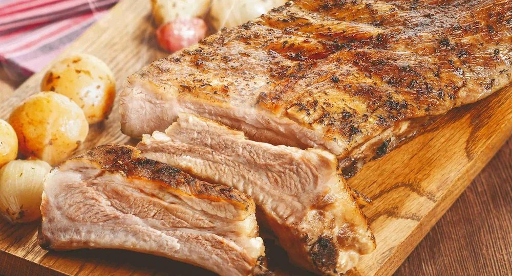 Uma suculenta peça de Costelinha Suína Frimesa assada, servida sobre uma tábua rústica de madeira. A carne apresenta uma crosta dourada e rica em temperos, com fatias recém-cortadas na parte da frente que revelam um interior claro e macio junto aos ossos. Acompanhando a costelinha, pequenas batatas e cebolas assadas estão espalhadas pela tábua, compondo um cenário quente e muito apetitoso com um pano listrado levemente desfocado ao fundo.