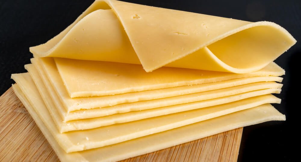 Pilha de fatias finas e amarelas de Queijo Prato Frimesa dispostas sobre uma tábua de madeira clara. A fatia do topo está levemente dobrada em forma de onda, destacando a textura suave e macia do queijo.