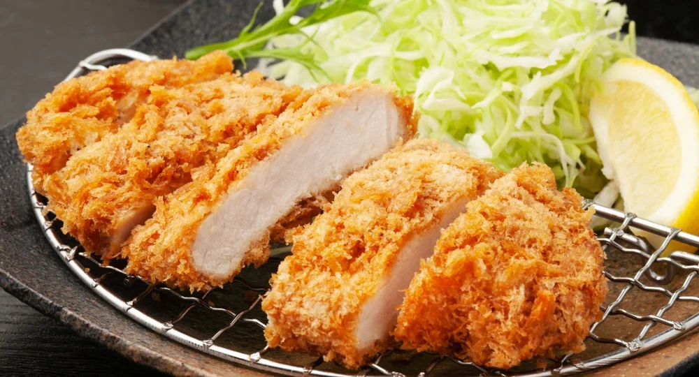 Frango empanado Frimesa perfeitamente fatiado, exibindo uma casquinha panko dourada e extremamente crocante por fora, com a carne macia e suculenta por dentro. As fatias descansam sobre uma pequena grade de metal para manter a crocância, acompanhadas de repolho verde desfiado e uma fatia de limão fresco ao fundo.