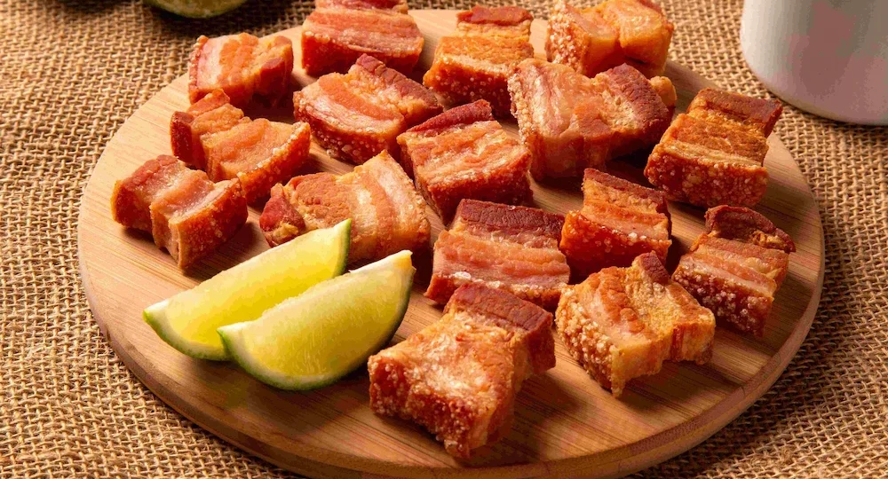 Vários cubos de panceta de porco fritos e pururucados servidos sobre uma tábua redonda de bambu. Os pedaços revelam camadas intercaladas de carne e gordura, cobertos por uma pele bem dourada, estufada em pequenas bolhas e com aspecto extremamente crocante. À esquerda na tábua, duas fatias de limão-tahiti fresco acompanham a porção. Ao fundo, a tábua está apoiada sobre um tecido rústico de juta em tons quentes.