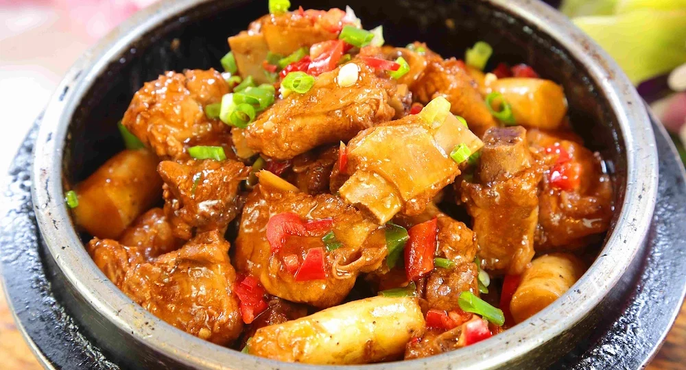 Uma apetitosa porção de Costelinha Suína Frimesa cortada em pequenos pedaços com osso, servida em uma panela rústica escura. A carne está perfeitamente cozida e envolta por um molho espesso, brilhante e caramelizado, misturada com pedaços claros de vegetais. O prato é finalizado com cebolinha verde fresca e pedacinhos de pimenta ou pimentão vermelho picado por cima, criando um contraste vibrante de cores e um visual extremamente suculento.
