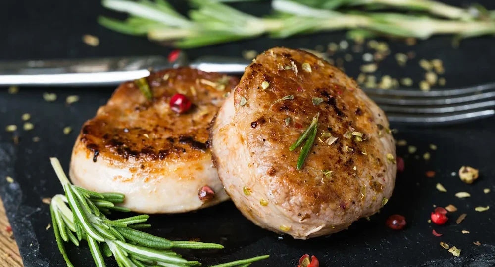 Dois suculentos medalhões de filé mignon suíno grelhados, com uma superfície bem dourada e apetitosa, servidos sobre uma base rústica de pedra escura. A carne está temperada com ervas secas e grãos de pimenta rosa, acompanhada por raminhos de alecrim fresco que dão um toque especial ao prato.