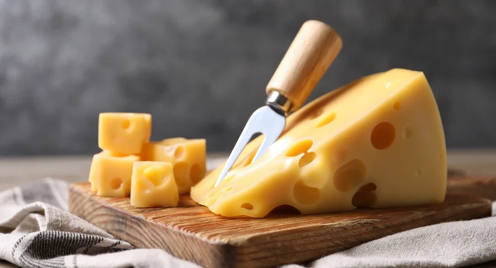 Na imagem, há um grande e apetitoso pedaço de queijo suiço Frimesa servido sobre uma tábua de madeira rústica. Há também uma faca própria para o queijo, com cabo de madeira, que está espetada no centro da fatia principal. Ao lado, repousam alguns cubos pequenos do queijo.