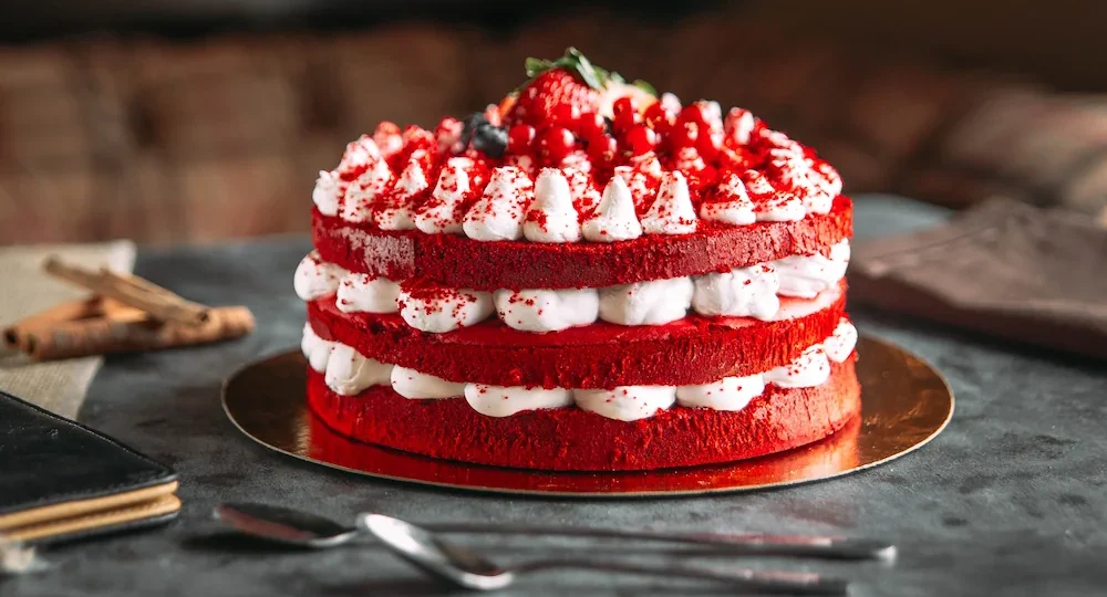 Um lindo bolo de três camadas com massa bem vermelha e bastante recheio de creme branco. O topo está decorado com o mesmo creme, pequenas migalhas de bolo e frutos vermelhos frescos, como morangos, no centro. O bolo está sobre uma base dourada, com duas colheres prateadas logo à frente, pronto para ser saboreado.