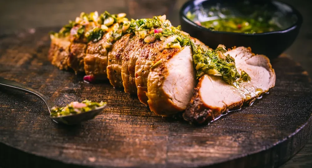 Peça de carne de porco Frimesa assada, cortada em fatias espessas, sobre uma tábua de corte em madeira rústica e escura. A carne tem uma crosta dourada e está generosamente coberta por um molho verde de ervas frescas picadas, semelhante a chimichurri. Em segundo plano, ligeiramente desfocada, encontra-se uma taça escura com mais molho. Em primeiro plano, à esquerda, vê-se uma colher também com um pouco do preparado de ervas.