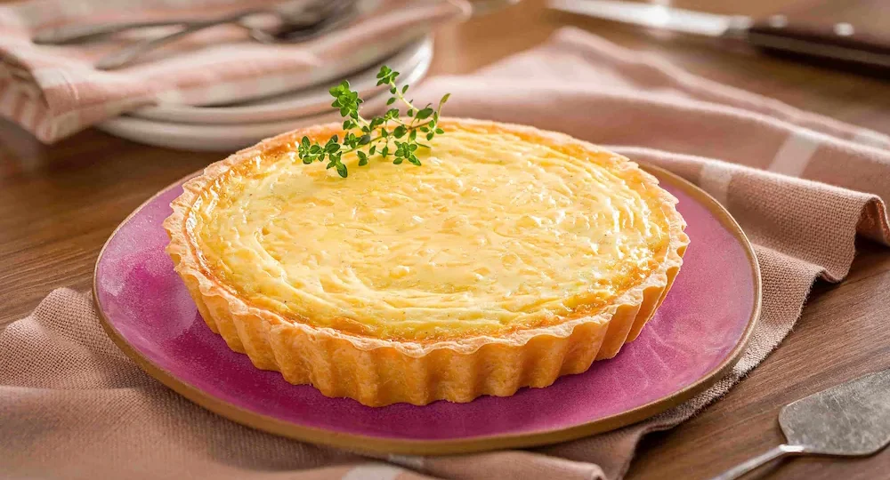 Uma quiche apetitosa, servida num prato roxo. A massa apresenta as tradicionais bordas onduladas e bem douradinhas, enquanto o recheio, de tom amarelinho e textura cremosa, é decorado com um ramo de ervas frescas. O prato está sobre um pano bege numa mesa de madeira, com alguns pratos e talheres ligeiramente desfocados ao fundo, criando um cenário que convida a uma refeição muito saborosa.