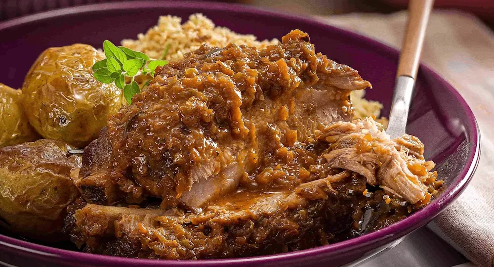 Um prato roxo fundo com uma suculenta e generosa porção de costelinha de porco suculenta. A carne mostra seu interior húmido e tenro, e está coberta por um molho espesso, rústico e bem apetitoso. No mesmo prato, também temos uma farofa e uma batata assada bem douradinha, além de pequeno ramo de ervas verdes. A luz acolhedora da fotografia realça a textura irresistível e caseira desta refeição.